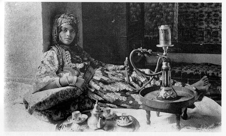 Czarno białe zdjęcie. Przedstawia młodą dziewczynę, która opiera się o ścianę i leży. Przed nią zastawa do herbaty, oraz stolik na którym stoi naczynie z wężami. Głębiej komoda i tkaniny. Dziewczyna ubrana jest w dekoracyjny strój, na głowie ma czepek z monetami, na rękach bransolety. Jest to odaliska paląca fajkę wodną. 