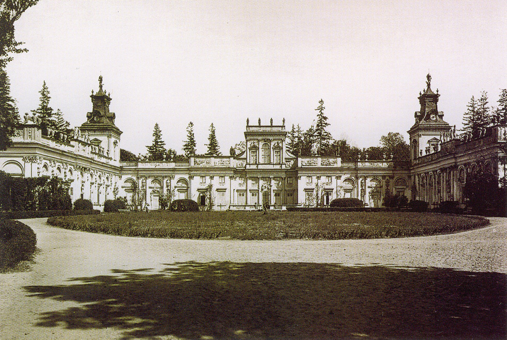 Dawna pocztówka przedstawiająca pałac w Wilanowie. Jest to czarno-biała fotografia, która pokazuje m.in. podjazd do pałacu, fragment zieleni i sam budynek w tle. 