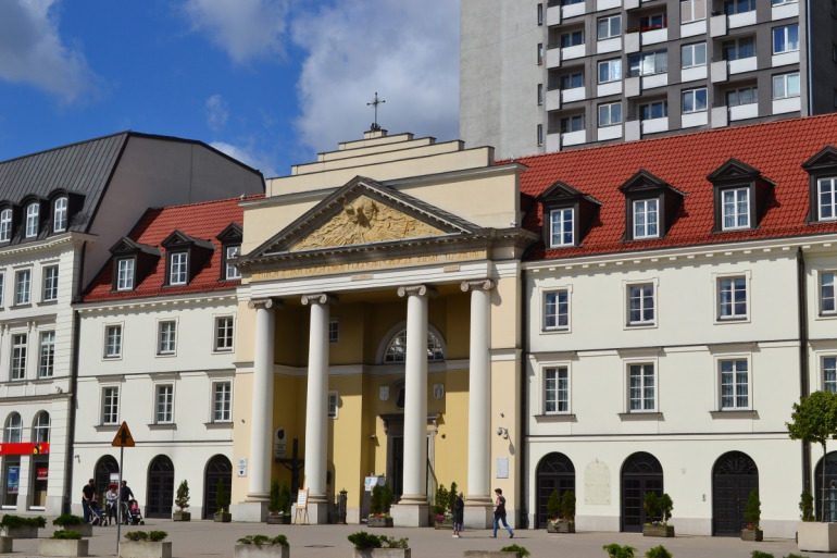 Kolorowe zdjęcie współczesne. Przedstawia widok na kamienice w fasadę kościoła. Jest to kościół  p.w. św. Brata Alberta i św. Andrzeja Apostoła w Warszawie. To tam znajduje się słynny obraz Matki Bożej, który według tradycji pochodzi z ołtarza polowego królów: Jana Kazimierza, Michała Korybuta Wiśniowieckiego a następnie Jana III Sobieskiego. 