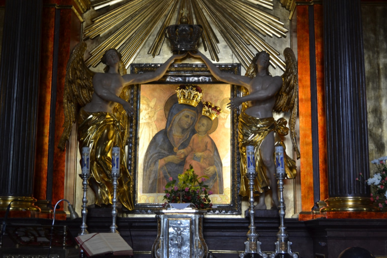 Obraz Matki Bożej Piaskowej. Maryja w granatowej sukni, podtrzymuje małego Jezusa w czerwonej szacie. W tle anioły. Maryja i dzieciątko mają na głowie złote korony. 15 sierpnia 1683 roku król Jan III Sobieski z rodziną odwiedził m.in. kościół ojców karmelitów na Piasku w Krakowie i łaskami słynący obraz Matki Bożej Piaskowej. Według legendy Jan III Sobieski i Maria Kazimiera właśnie przed tym wizerunkiem przyrzekli ufundować nowe kościoły – król dla ojców kapucynów, a królowa dla sióstr sakramentek. 