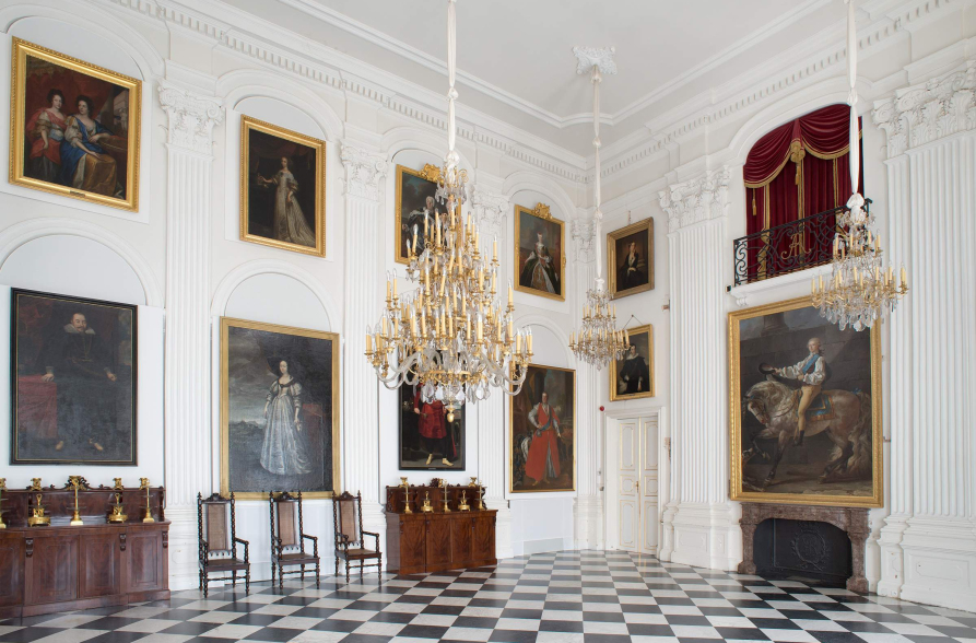 Sala pałacowa w kolorze białym ze złotym żyrandolem. Posadzka w czarno-białą szachownicę. Na ścianach wiszą obrazy w złotych ramach. Po prawej stronie stoi fortepian, po lewej stronie rząd krzeseł.